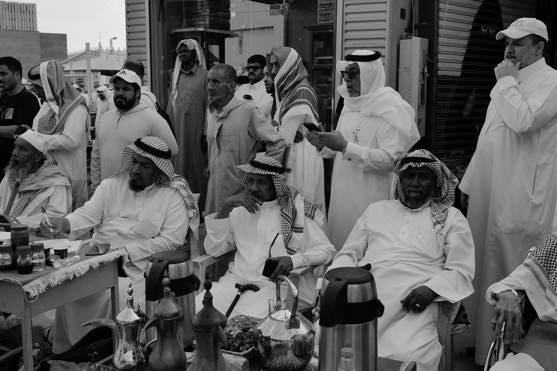 A group of Middle Eastern men, many wearing traditional white robes and keffiyeh head coverings, gathered outdoors, some seated at a table with bottles and equipment, others standing or using phones, with a modern building in the background.