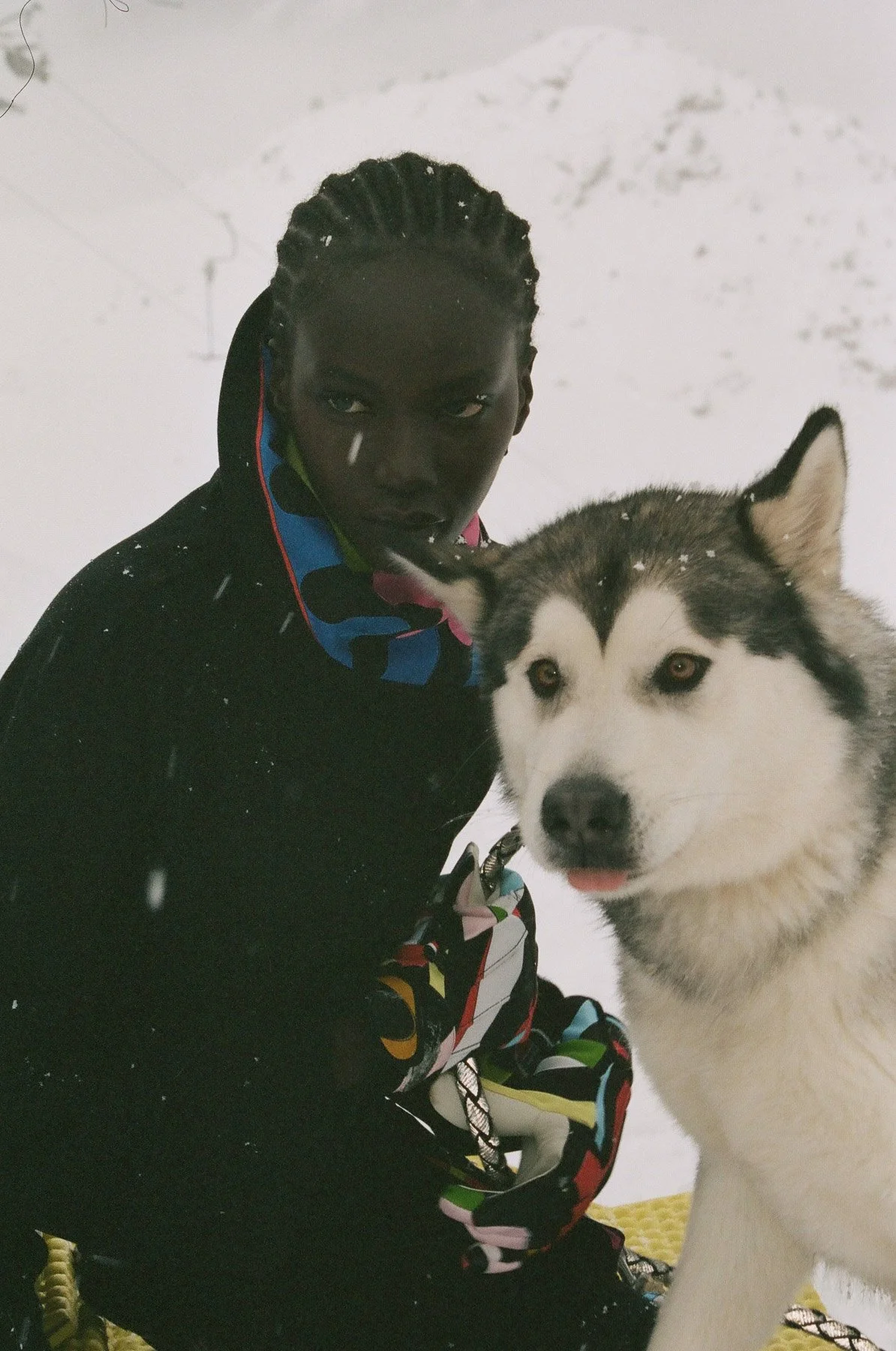 A person with braided hair and dark skin in a black coat with a colorful pattern takes a selfie with a Siberian Husky dog in a snowy environment.