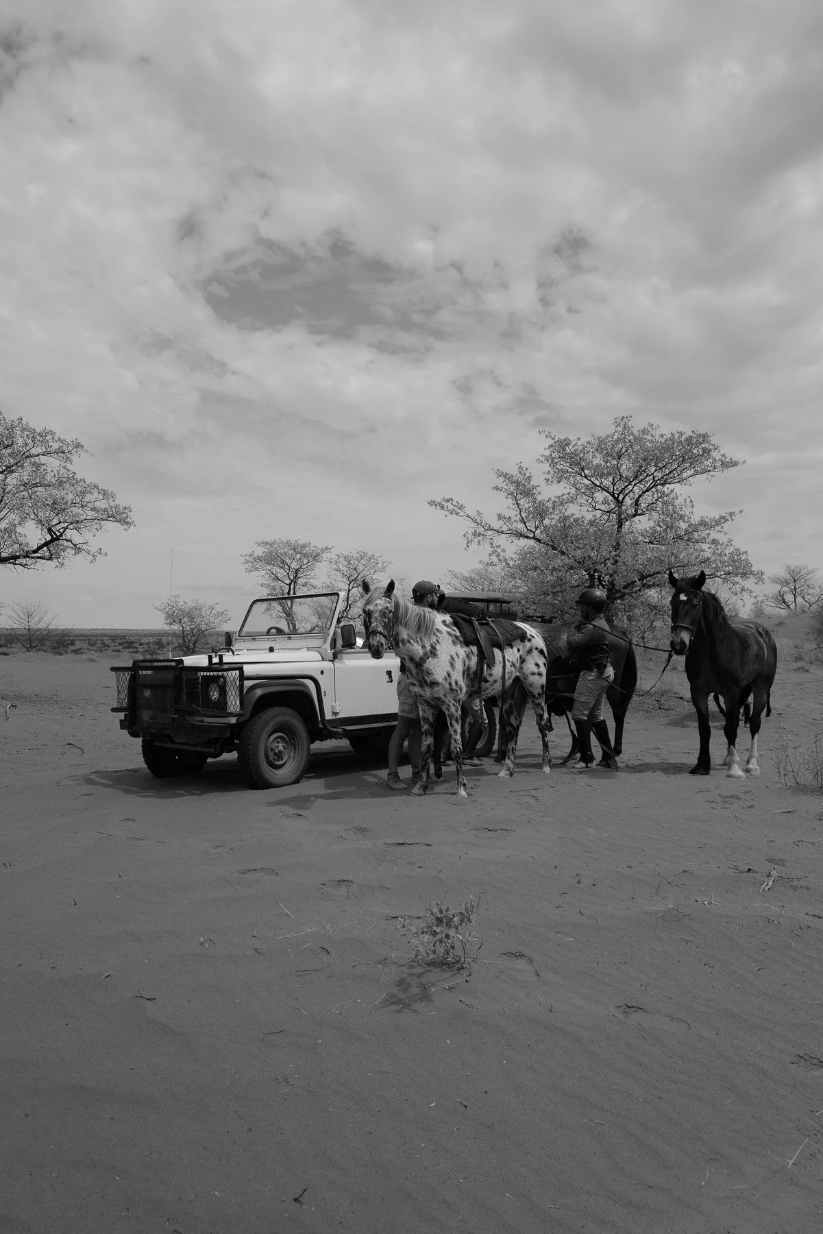 Two people on horseback with a safari vehicle in a dry, open landscape with scattered trees and cloudy sky.