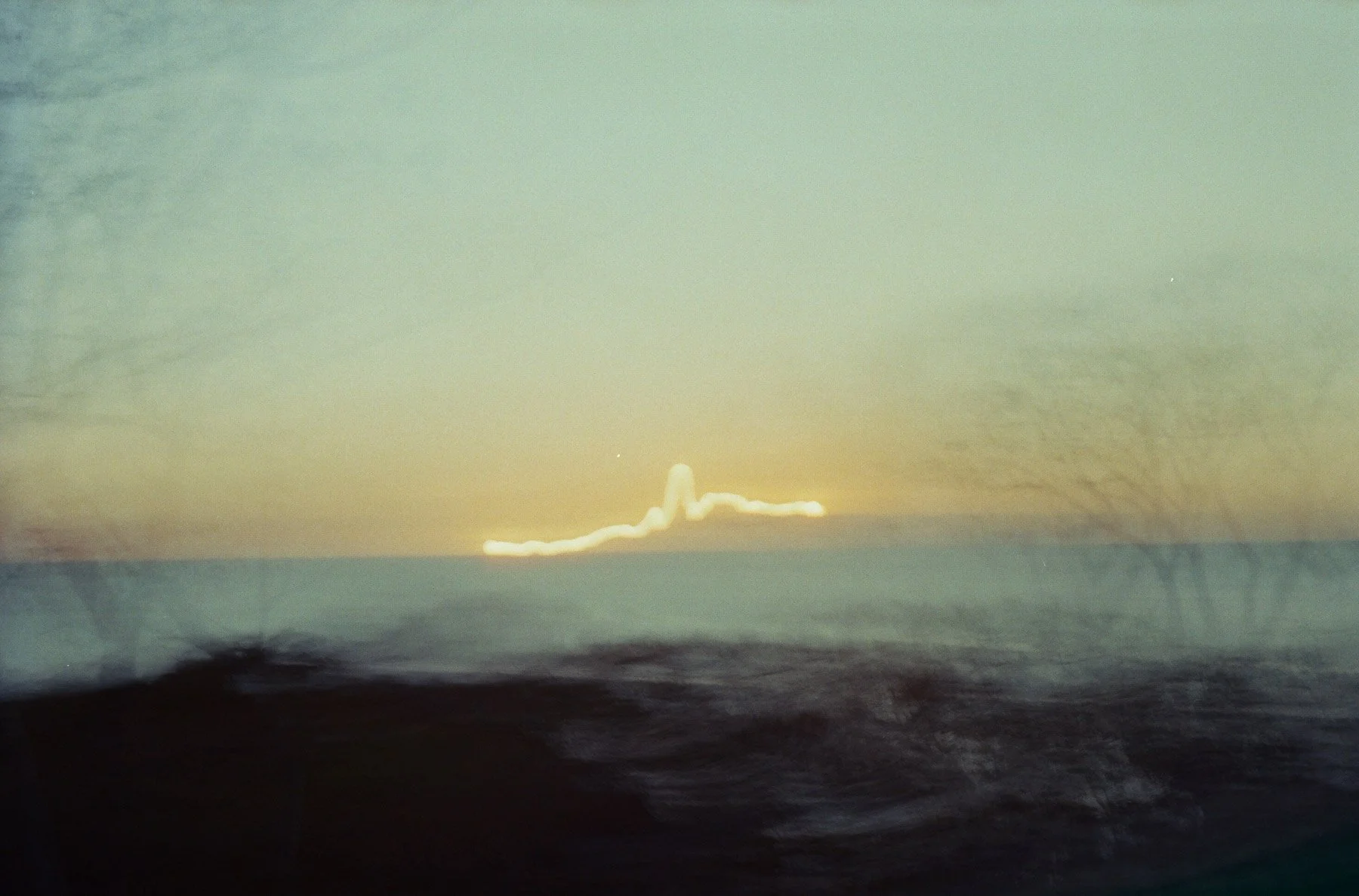 Photo of a distant explosion or lightning in the sky near the horizon, with a mix of yellow, green, and blue colors in the sky and dark landscape in the foreground.
