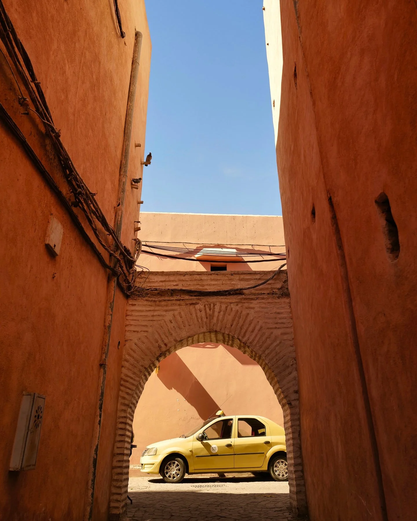 A yellow taxi parked under a brick archway in a narrow alley with orange walls and a clear blue sky.
