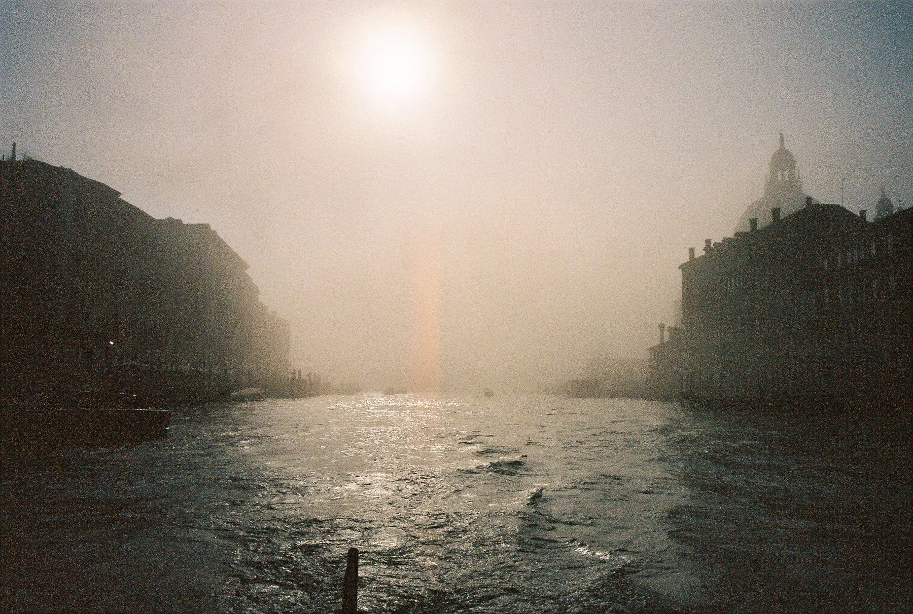 A foggy river scene at sunset with silhouettes of historic buildings and a dome-shaped structure in the distance.