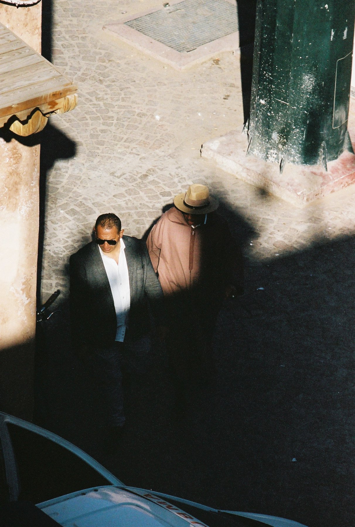 Two men walking on a cobblestone street, one dressed in a suit with sunglasses, and the other in a tan hat and coat. They are near a large green pole and a corner with a wooden structure.