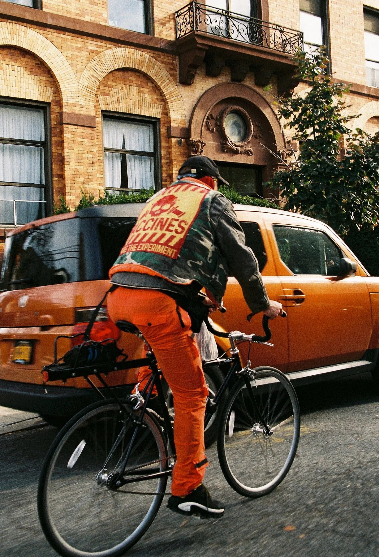 A person wearing an orange outfit and a jacket with a large patch reading 'Vaccines Save Lives' rides a black bicycle past a parked orange car in front of a brick building with arched windows and a decorative balcony.
