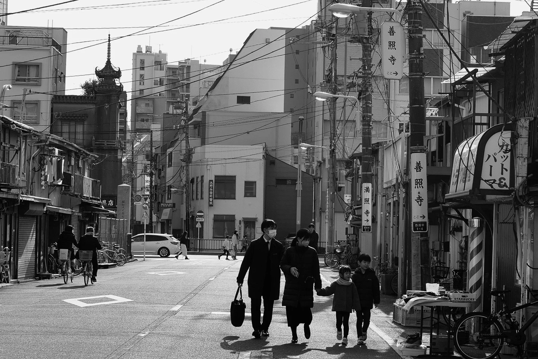 Street scene in Japan with pedestrians, bicycles, and shops in a neighborhood. Power lines are overhead, traditional and modern buildings line the streets, and some people wear masks.