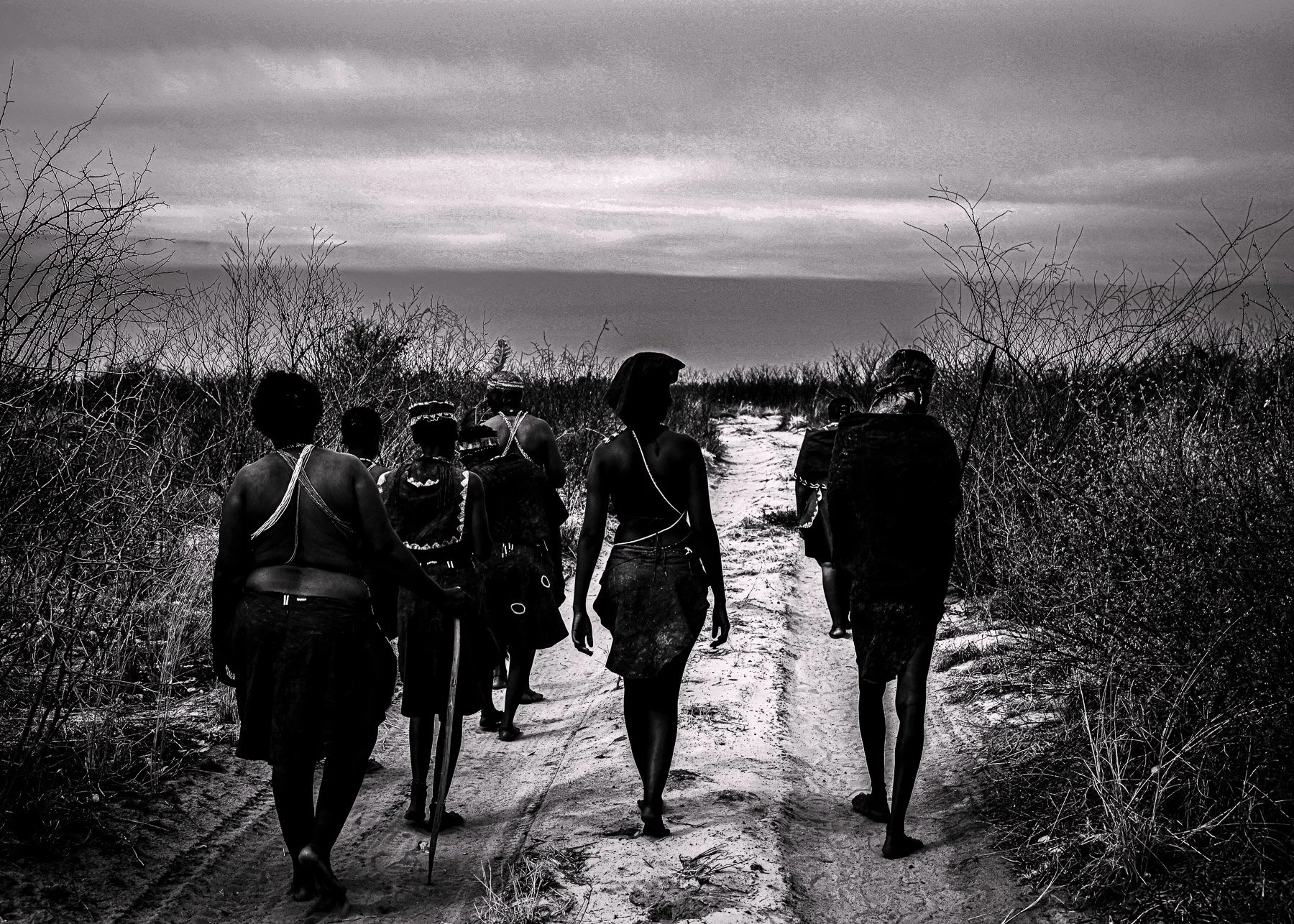 A group of indigenous people walking along a dirt trail through a dry, bushy landscape under a cloudy sky.