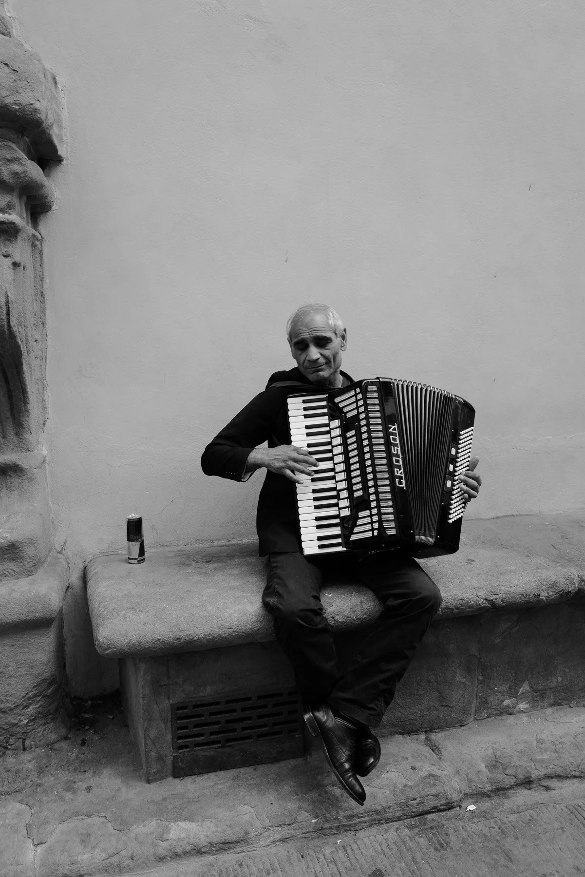 A man with gray hair sits on a stone bench, playing an accordion. He is dressed in dark clothing and looks down at his instrument with a contemplative expression. The background features a plain wall and a stone sculpture on the left, with a small ca