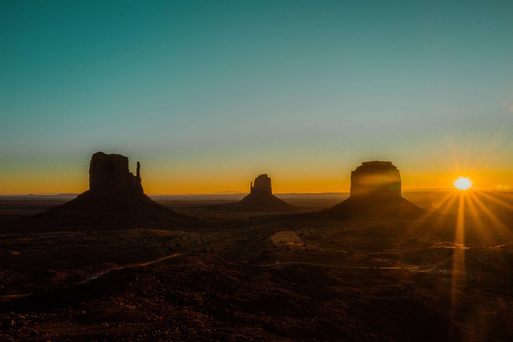 Sunset over Monument Valley with three large rock formations silhouetted against an orange and teal sky.