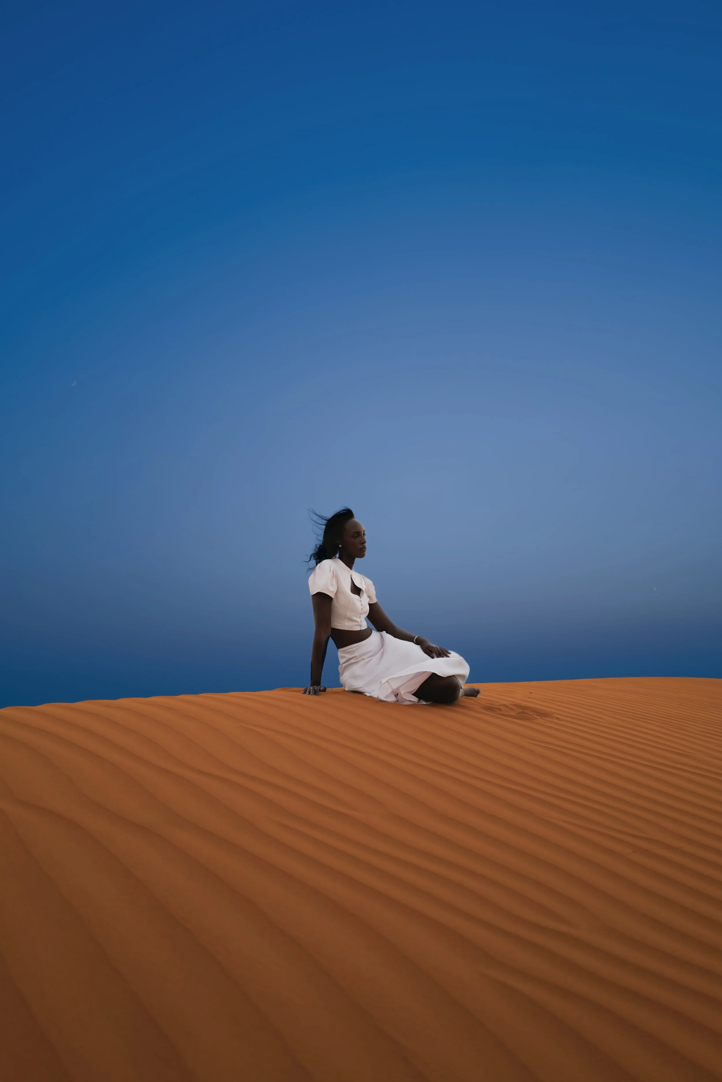 A woman sitting on orange sand dunes under a clear blue sky.