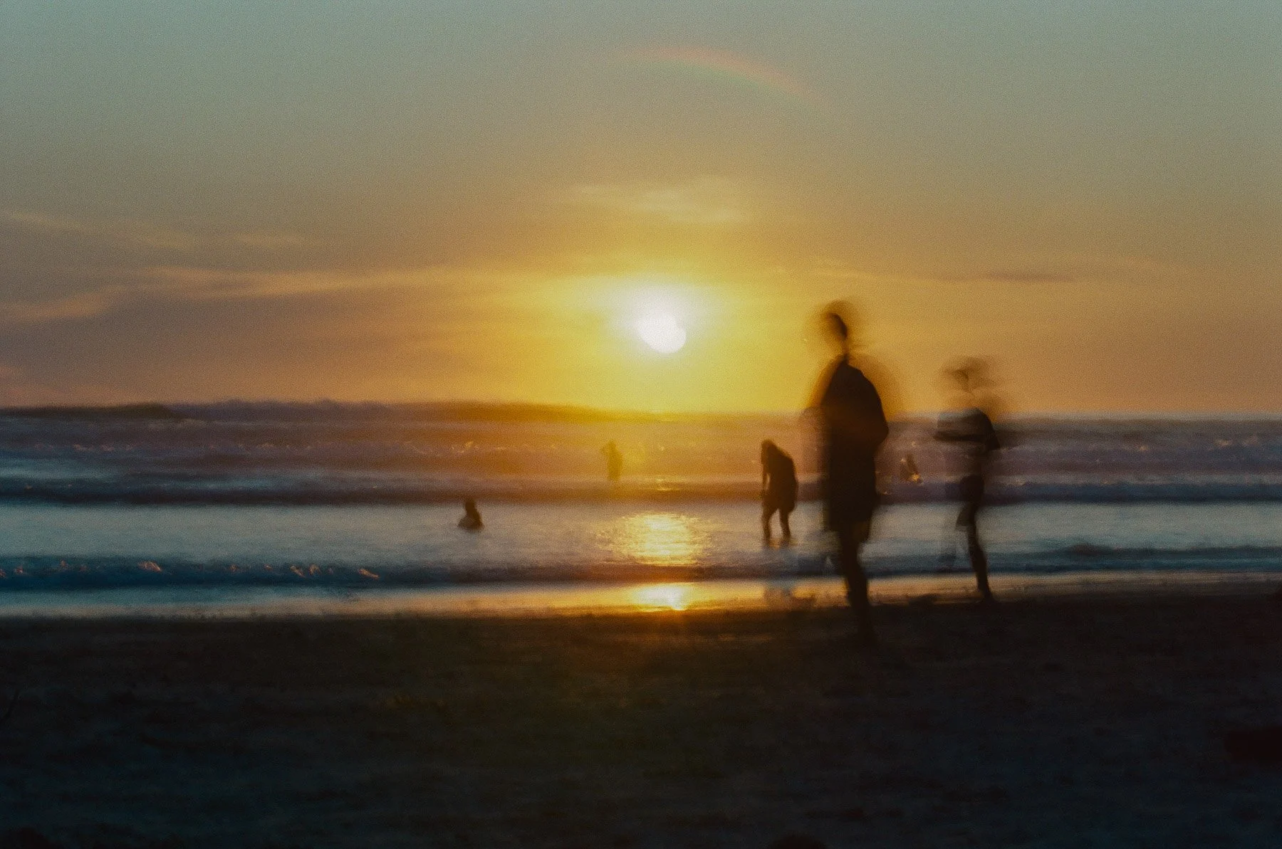 People walking and playing on the beach during sunset, with a rainbow visible in the sky.