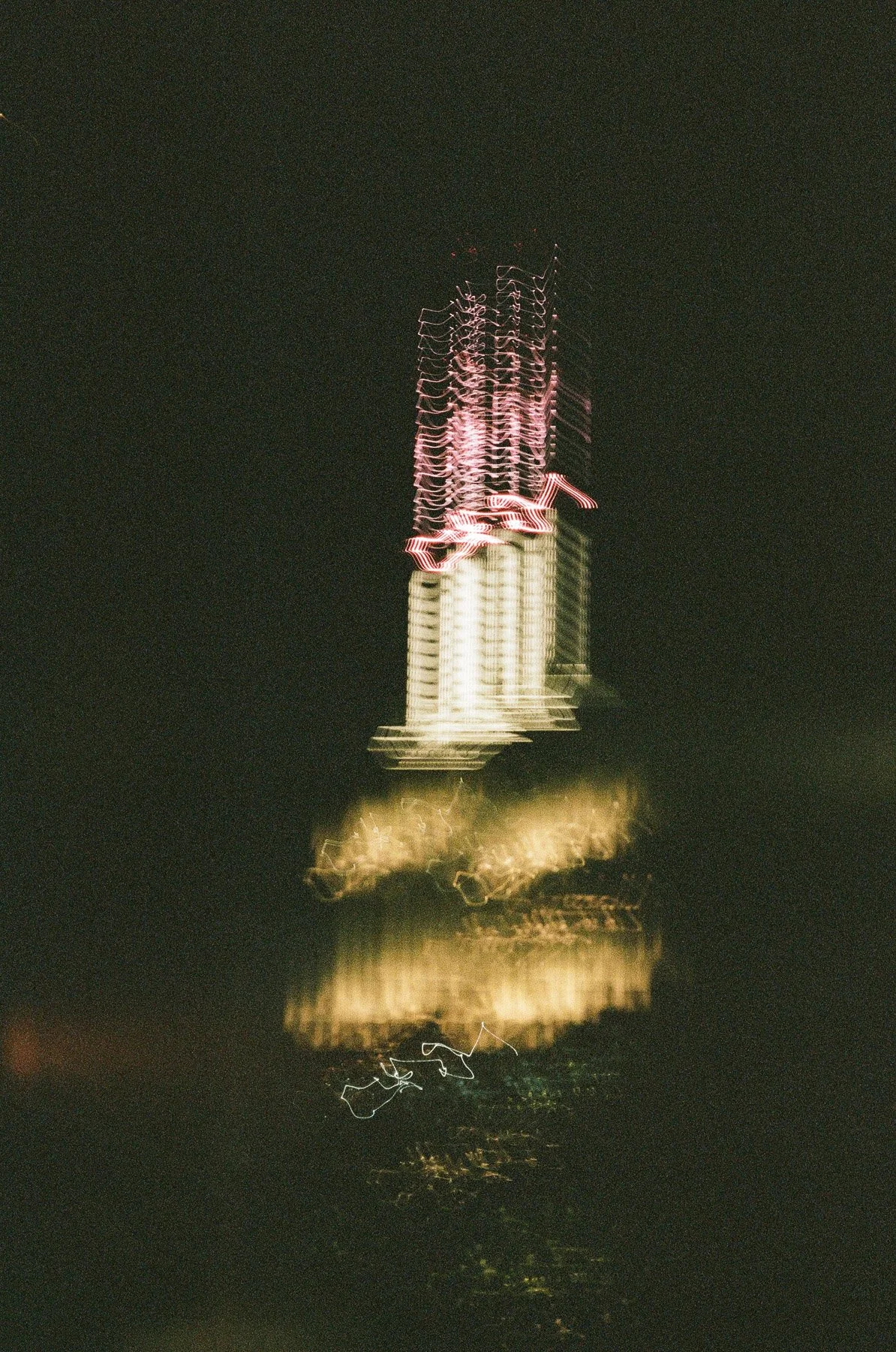 Blurred photo of a tall building, illuminated with pink and yellow lights, reflecting in water at night.