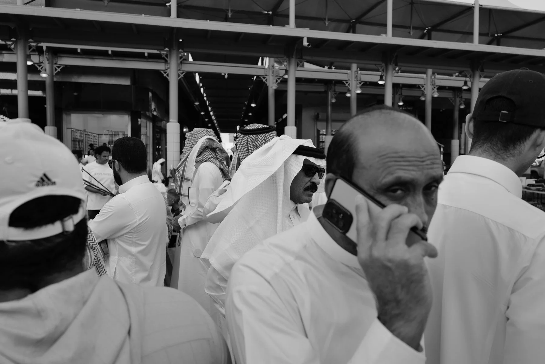 A black and white photo of Middle Eastern men in traditional attire, some wearing keffiyehs, gathered outdoors near a modern building with a metal roof structure. San Francisco man with sunglasses is on the phone in the foreground.