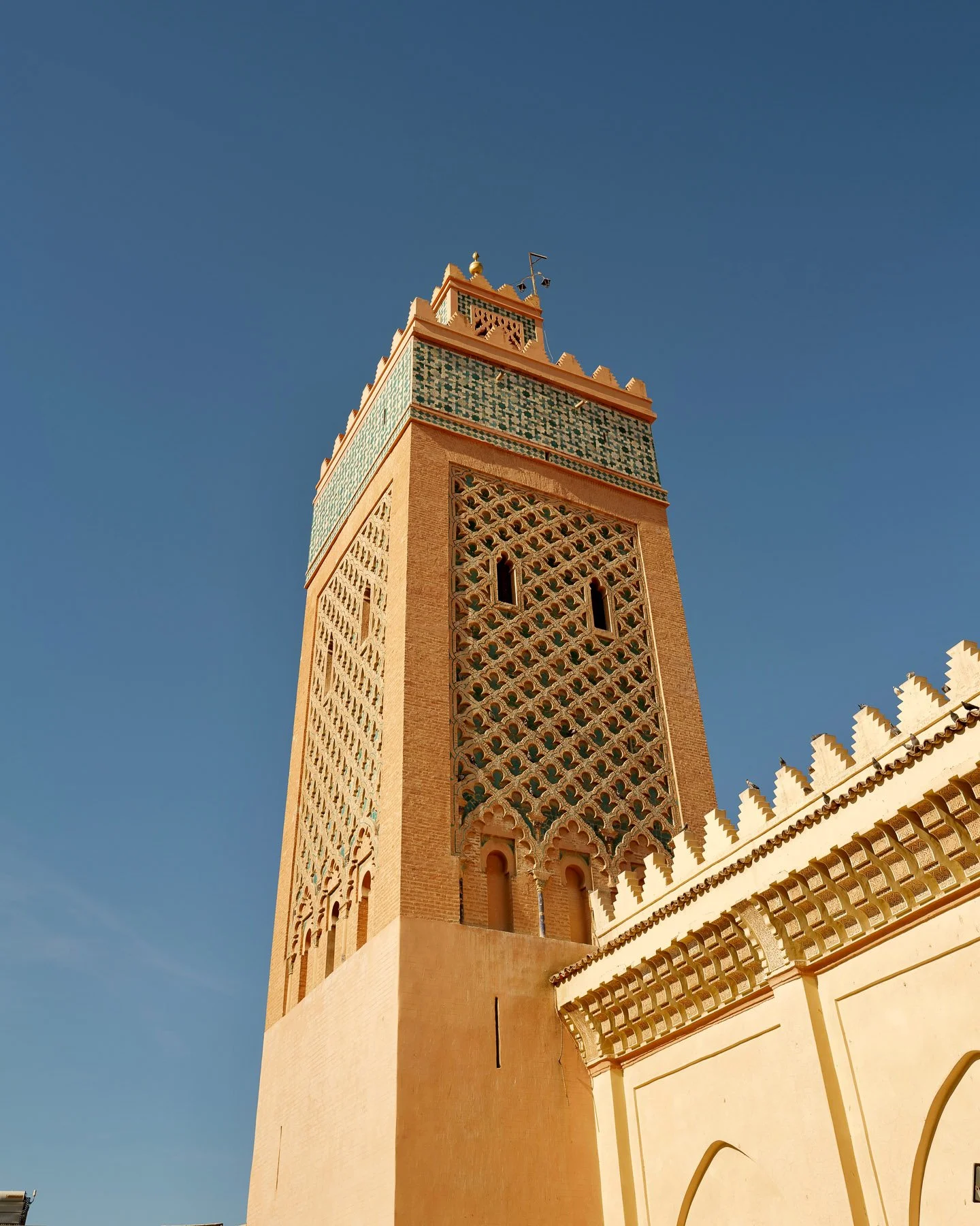 A tall mosque minaret with intricate tile work near the top, set against a clear blue sky.