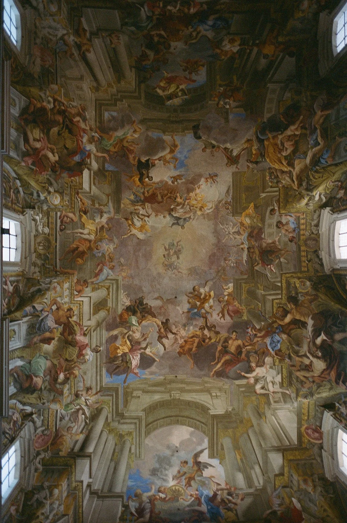 A grand baroque-style ceiling fresco depicting numerous angels, saints, and mythological figures painted in vibrant colors, with elaborate gold and white architectural details surrounding the artwork.
