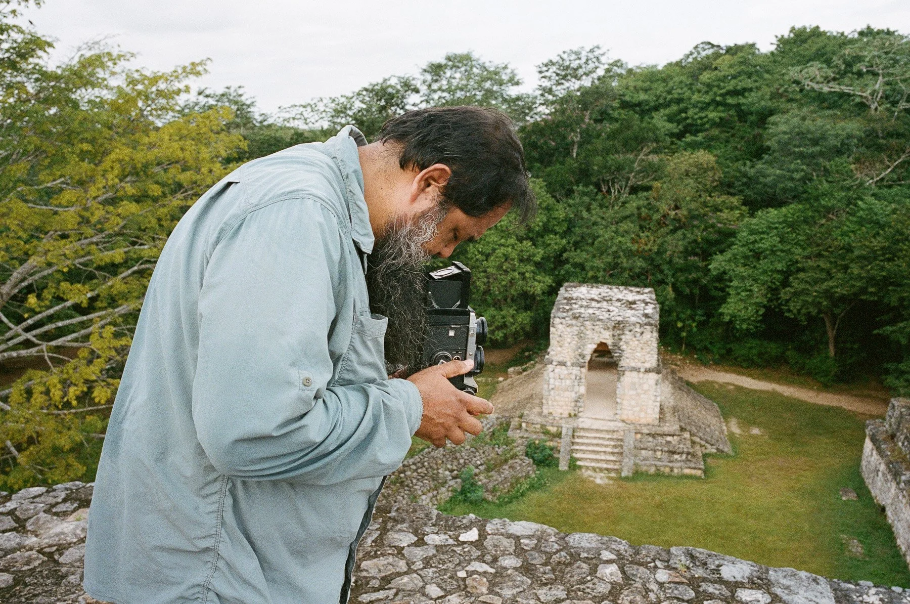 A man with a beard wearing a light blue shirt holding a vintage camera, standing near ancient ruins surrounded by green trees.