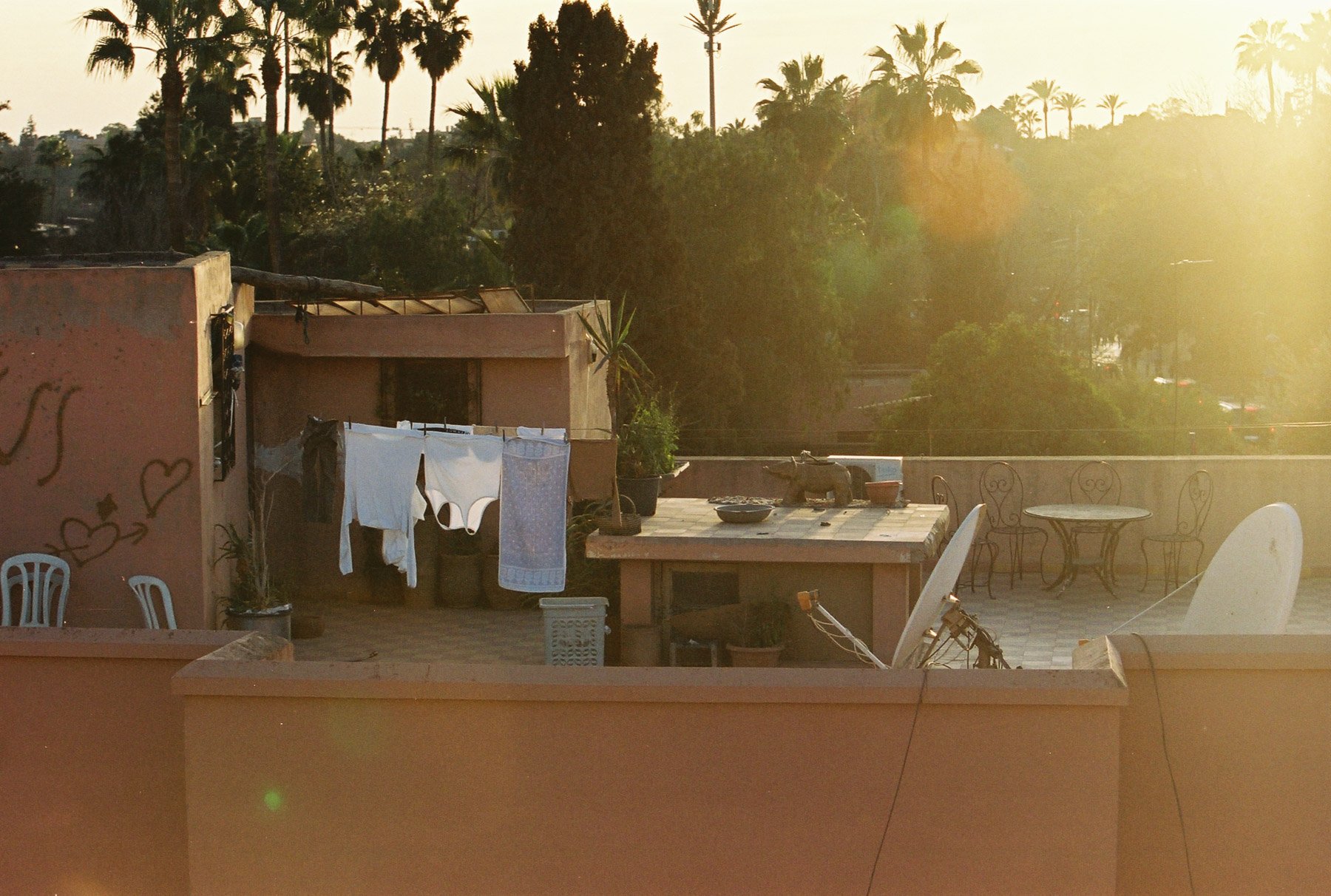 Rooftop patio with laundry hanging to dry, table with dishes and bowls, chairs, plants, and a view of palm trees and other greenery in the background during sunset.