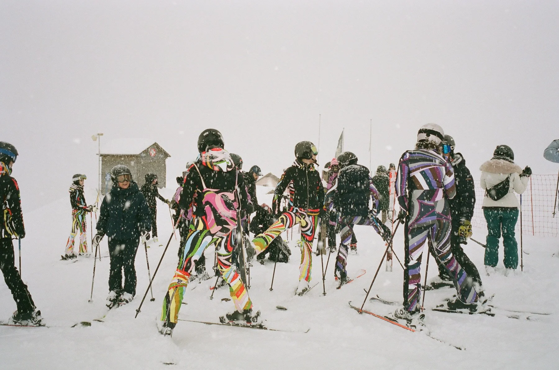 Group of people in colorful ski gear standing on snowy mountain during snowfall, preparing for skiing.