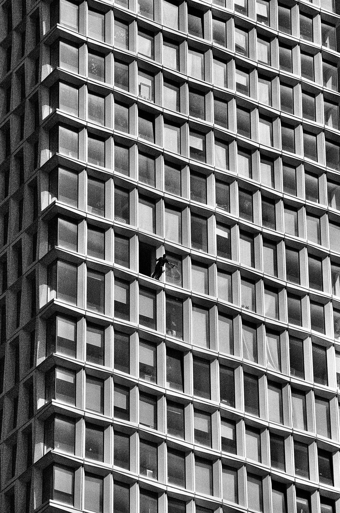 A black and white photo of a high-rise building with numerous square windows. A window cleaner is seen mid-action, hanging from a harness on one of the windows.