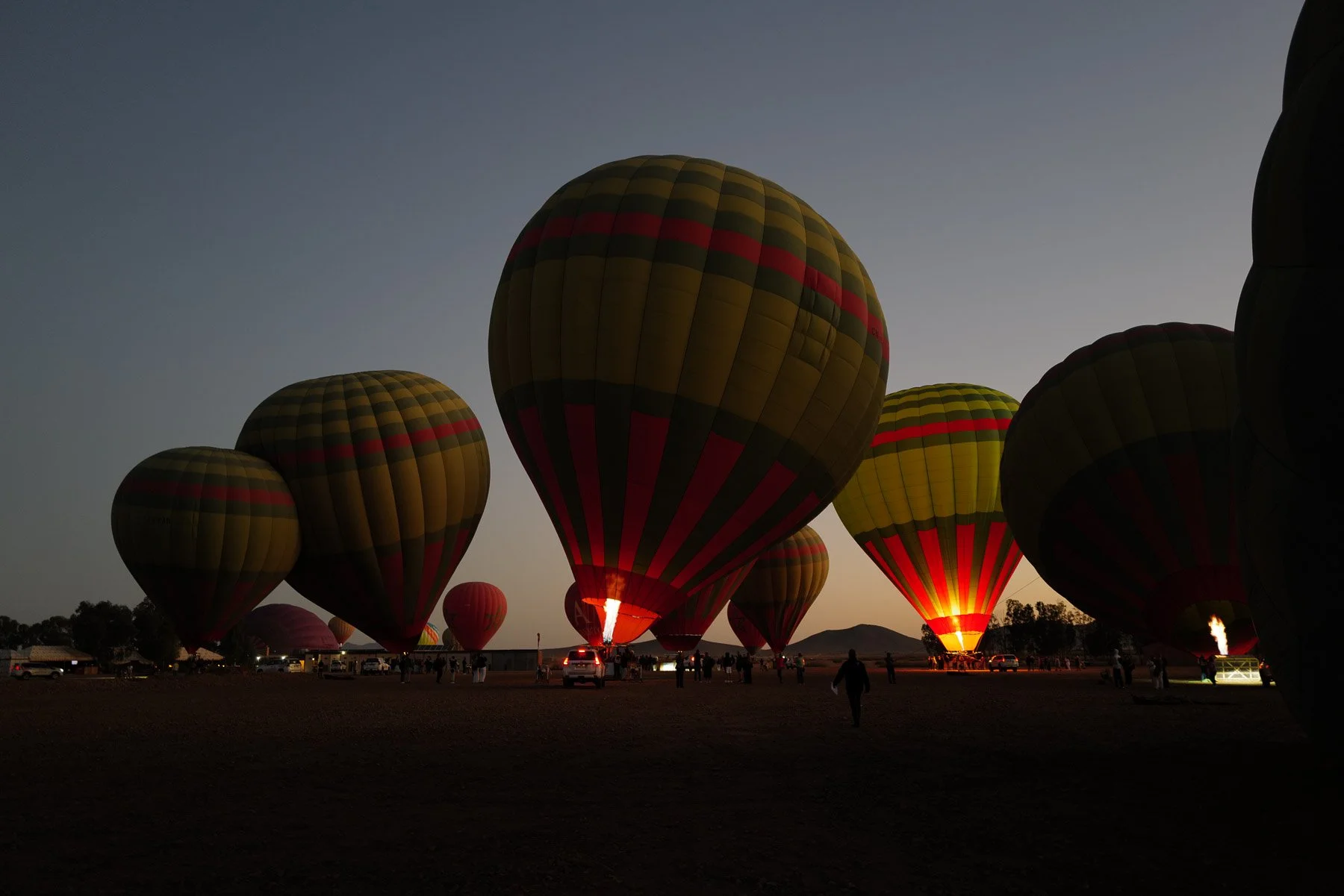 Hot air balloons being inflated and prepared for flight at dusk, with some balloons partially airborne and flames visible inside the balloons.