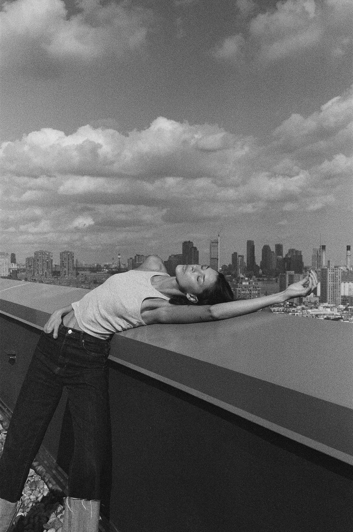 A woman lying on her back on a rooftop ledge with a city skyline in the background, stretching her arm out and looking upward.
