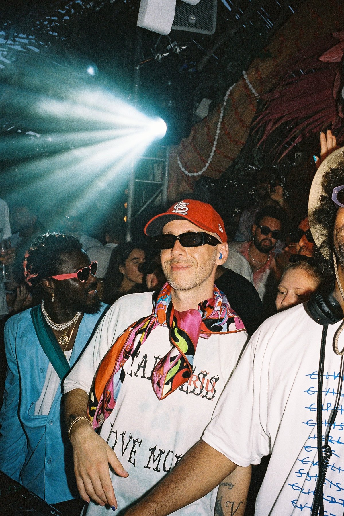 A group of people at a party or event, with colorful lighting and a festive atmosphere. The central figure is a man with glasses, a red cap, and a colorful scarf, standing near a DJ setup. Surrounding him are others wearing sunglasses and fashionable