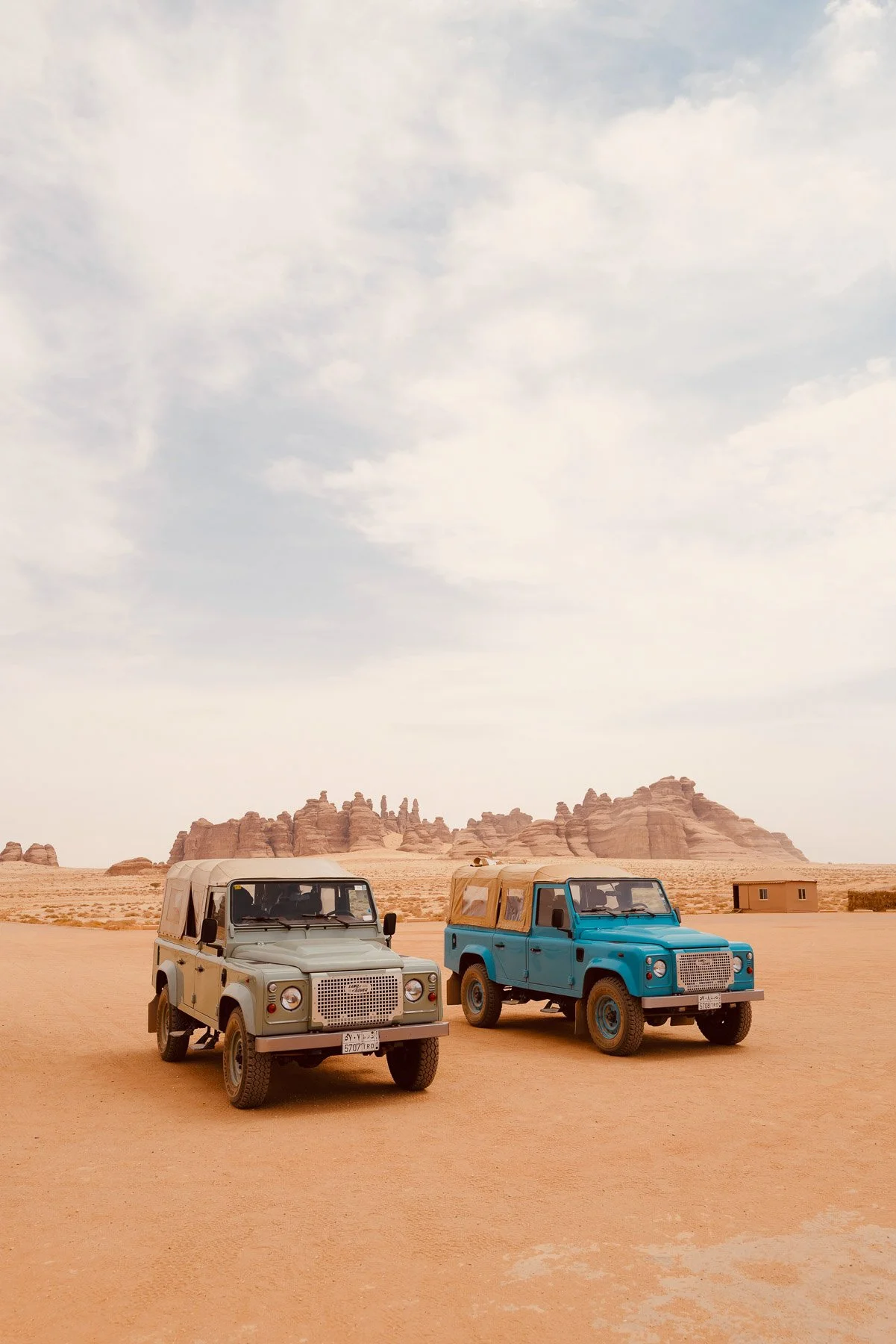 Two vintage Land Rover vehicles parked on a desert landscape with rock formations in the background under a cloudy sky.