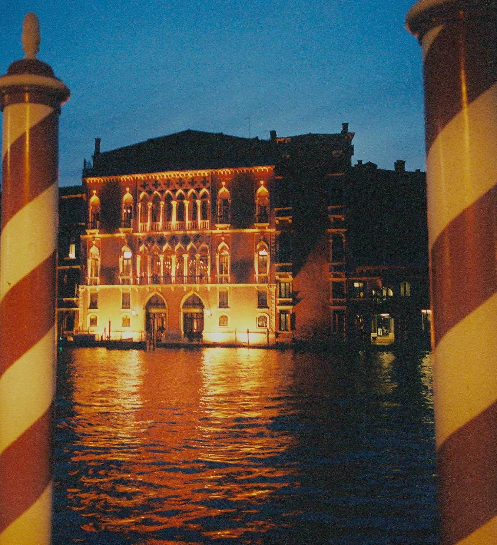 Nighttime view of a historic brick building with pointed windows and arches, illuminated with warm orange lights, reflecting on the water below, framed by striped yellow and brown walls.