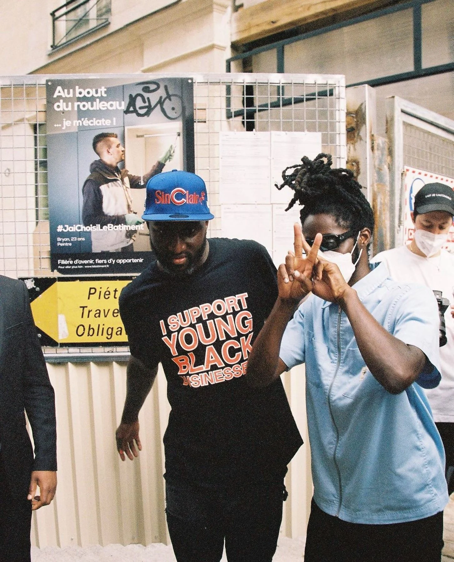 Two young men wearing face masks posing for the camera, one is making a peace sign, and the other is wearing a black T-shirt with the words 'Support Young Black Businesses' in bold, in front of a sign with images and text, including a poster with a y