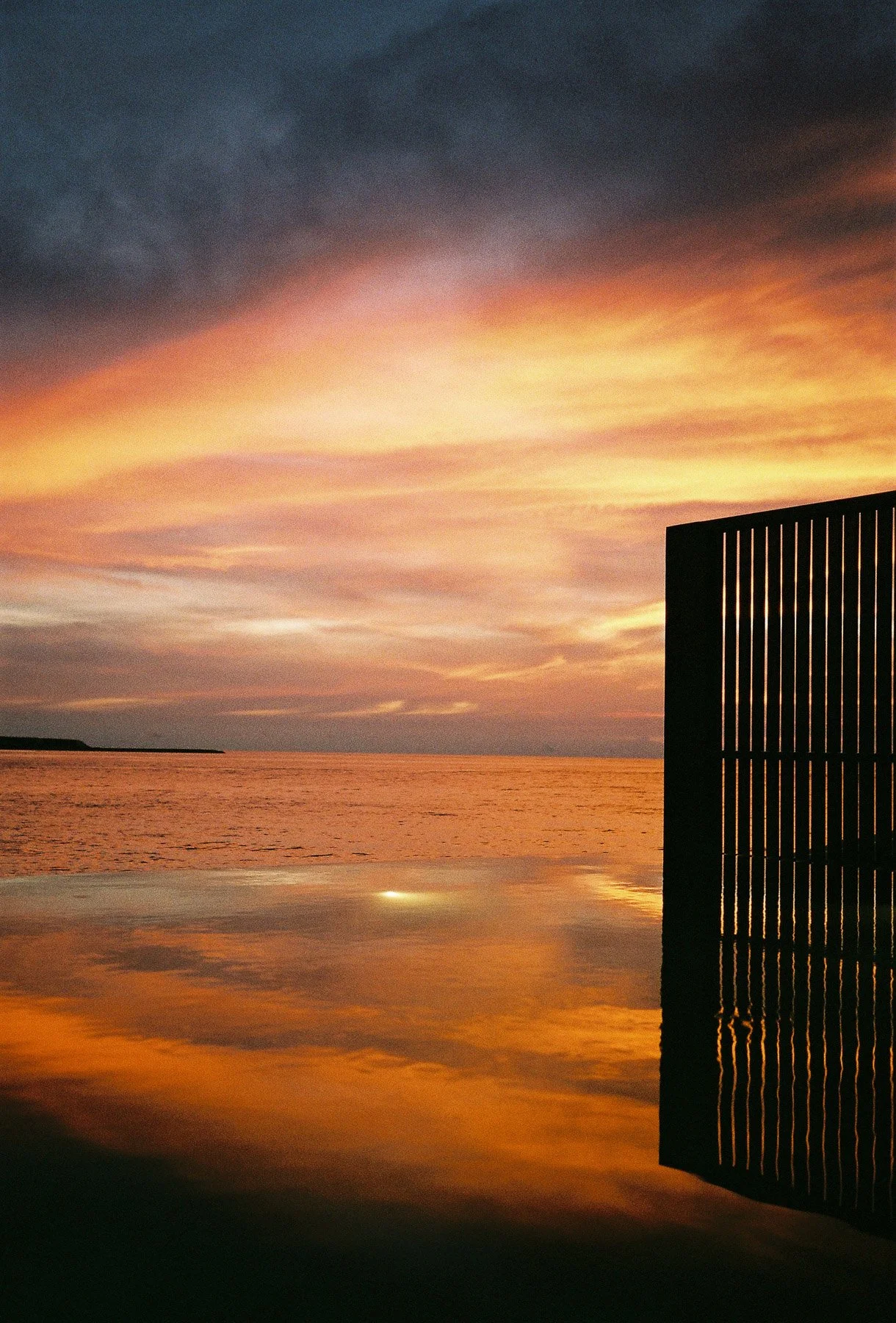 Sunset over a body of water with a black slatted structure on the right side and the water reflecting the colorful sky.