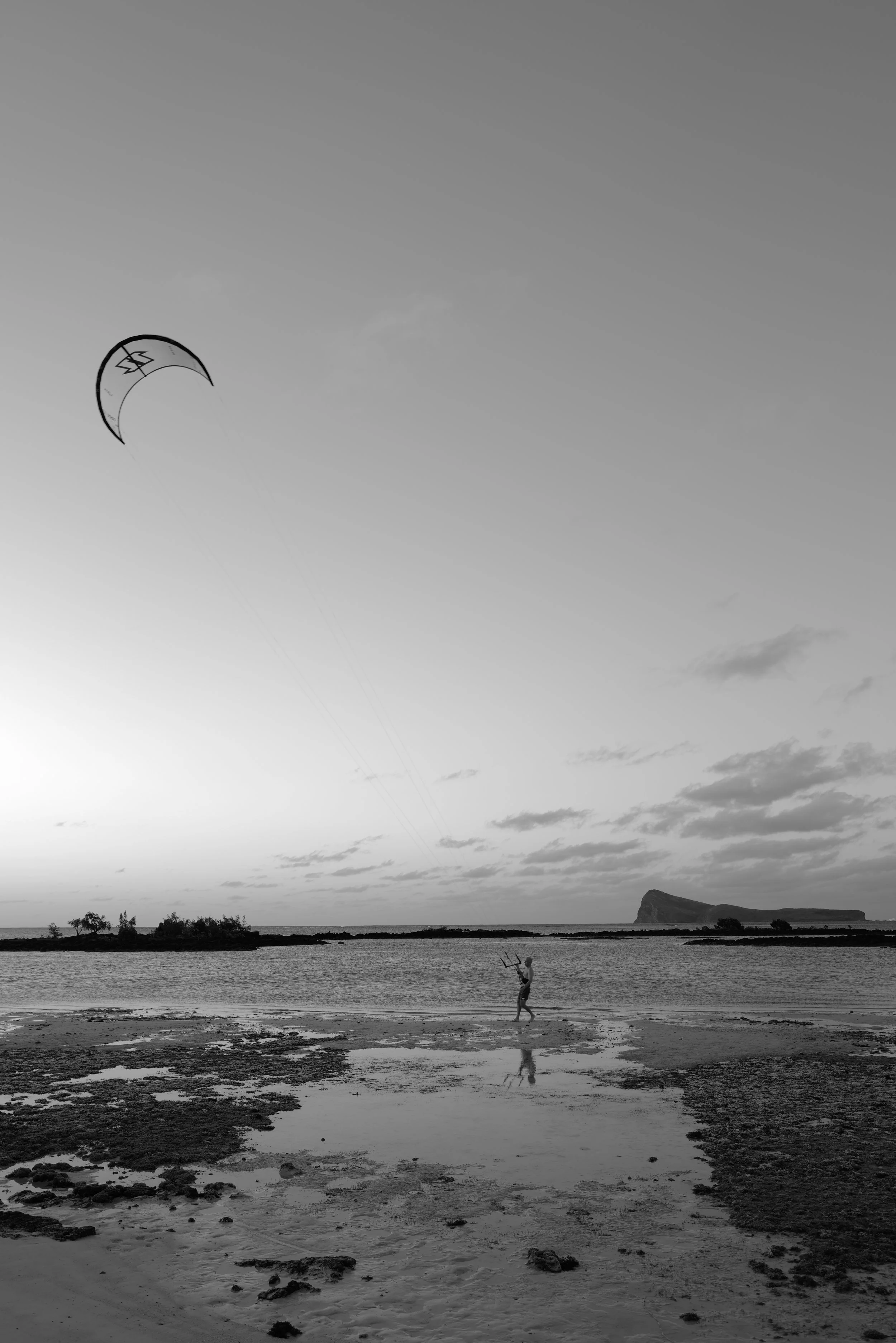 A person flying a kite on a beach at sunset, with water, rocky formations, and cloudy sky in the background, in black and white.