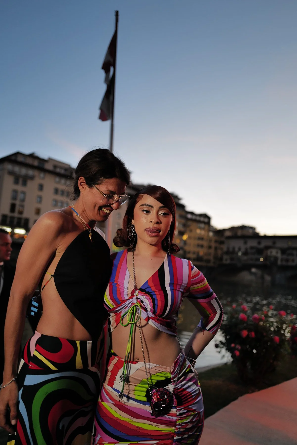 Two women standing outdoors near water with buildings in the background during sunset. One woman is wearing a black sleeveless top and colorful skirt, and the other woman is wearing a colorful, striped, and ruched dress, with accessories including la