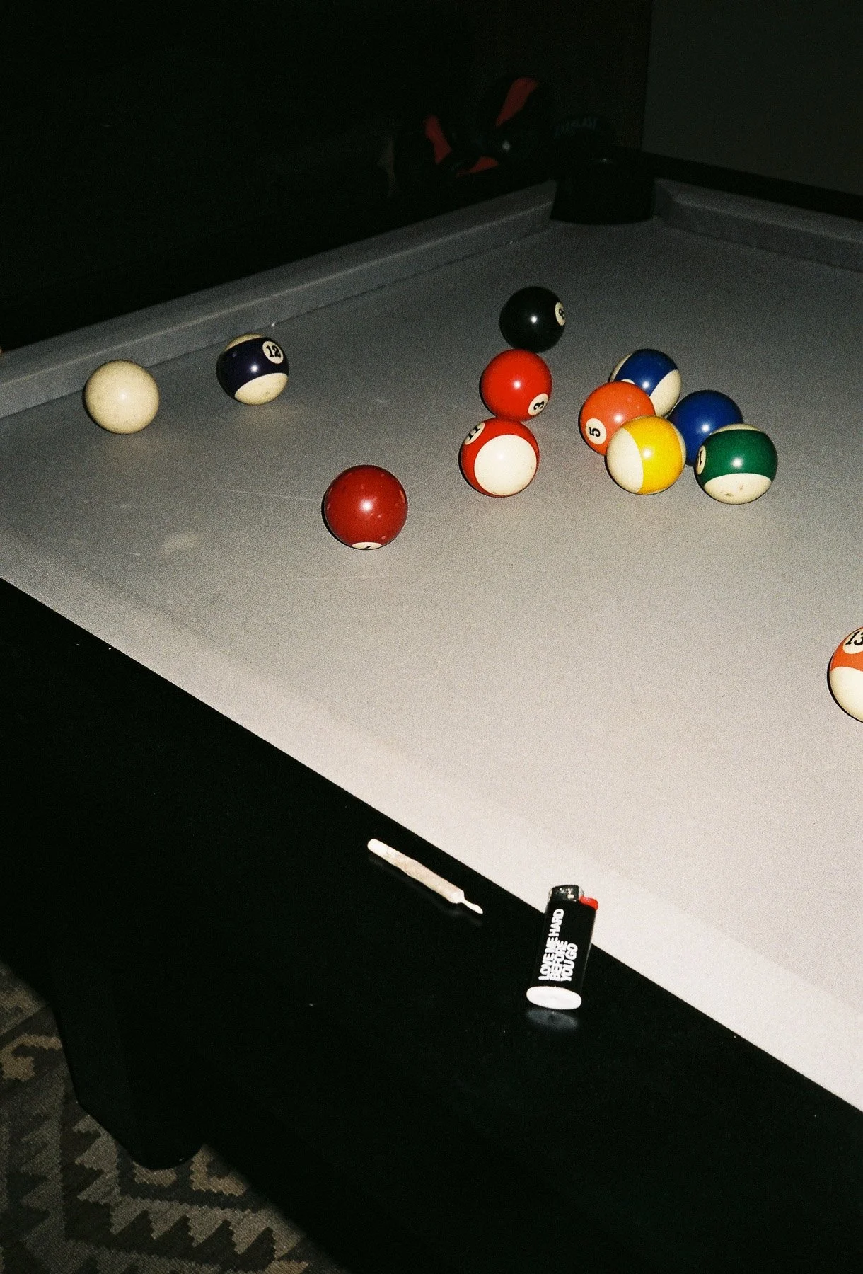 Pool table with billiard balls racked and some balls scattered, cue stick resting on edge, dark background.