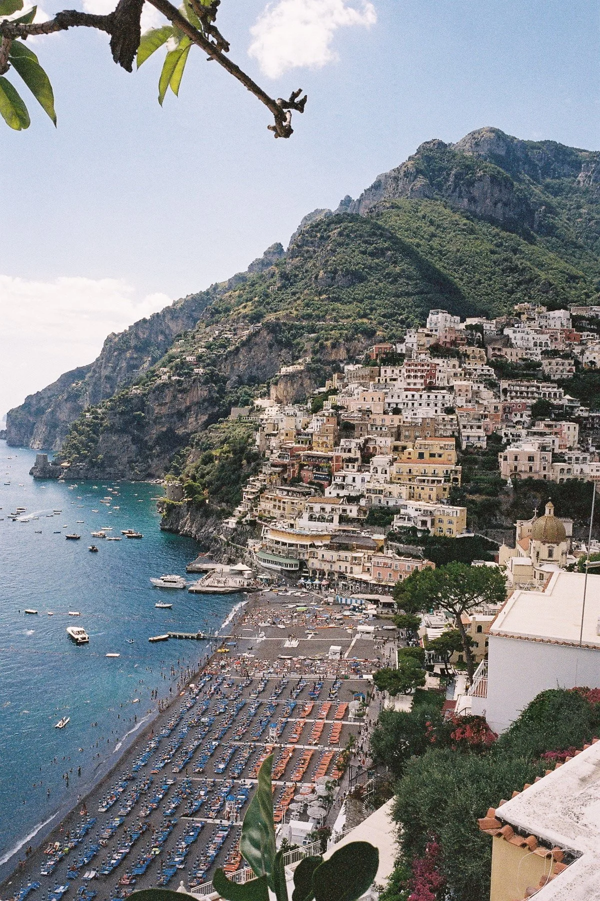 A coastal town built on steep hillsides with colorful buildings, a beach with umbrellas and lounge chairs, and boats on the water, with mountains in the background.
