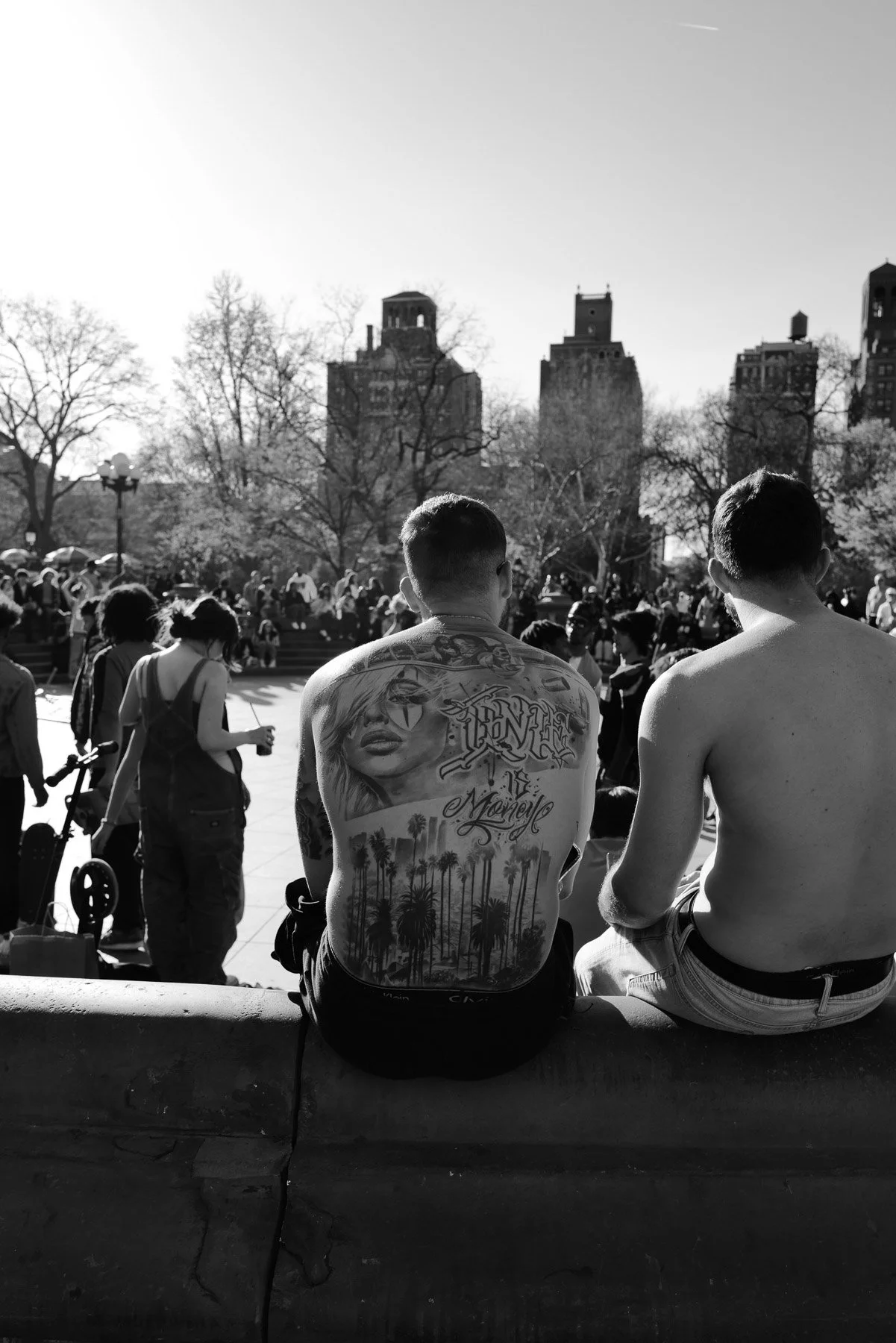 Two shirtless men sitting on a ledge with their backs to the camera in a park or square, surrounded by other people and trees, with city buildings in the background.