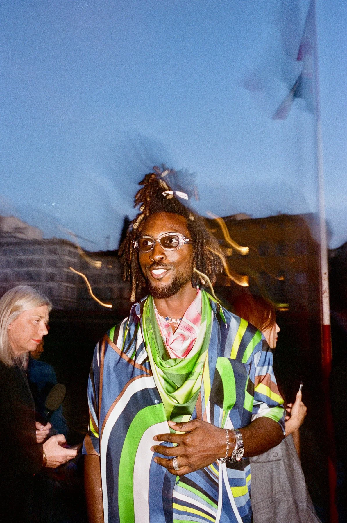 A man with dreadlocks, glasses, and a beard smiling, wearing a colorful abstract shirt with a green scarf and accessories, standing outdoors at dusk with other people in the background.
