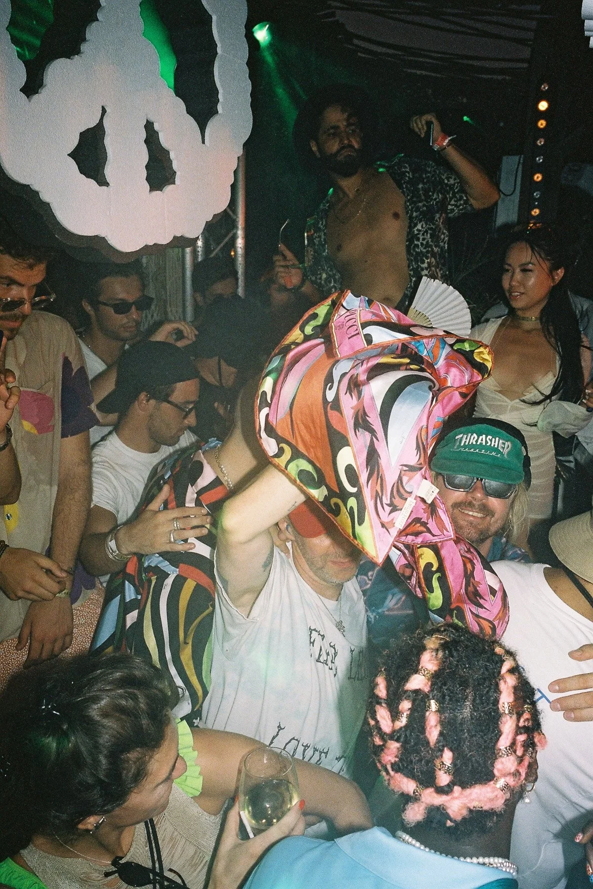 A group of people at a party or music event, some wearing sunglasses and casual clothing, with a man in the background shirtless and wearing a hat, surrounded by colorful decorations and a dark atmosphere with stage lighting.