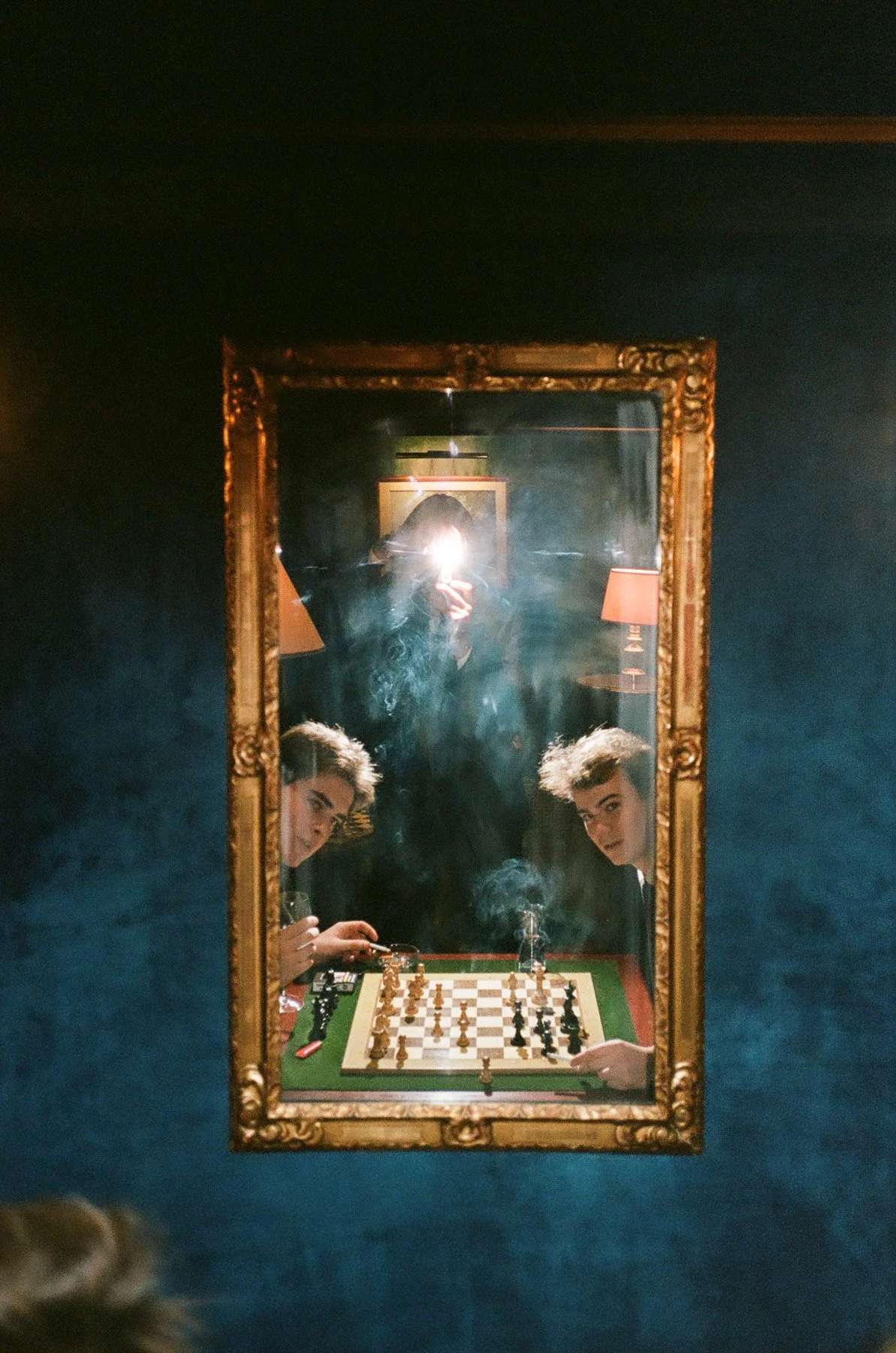 A person taking a selfie in a mirror with two young women playing chess at a table, with lamps and framed art in the background.