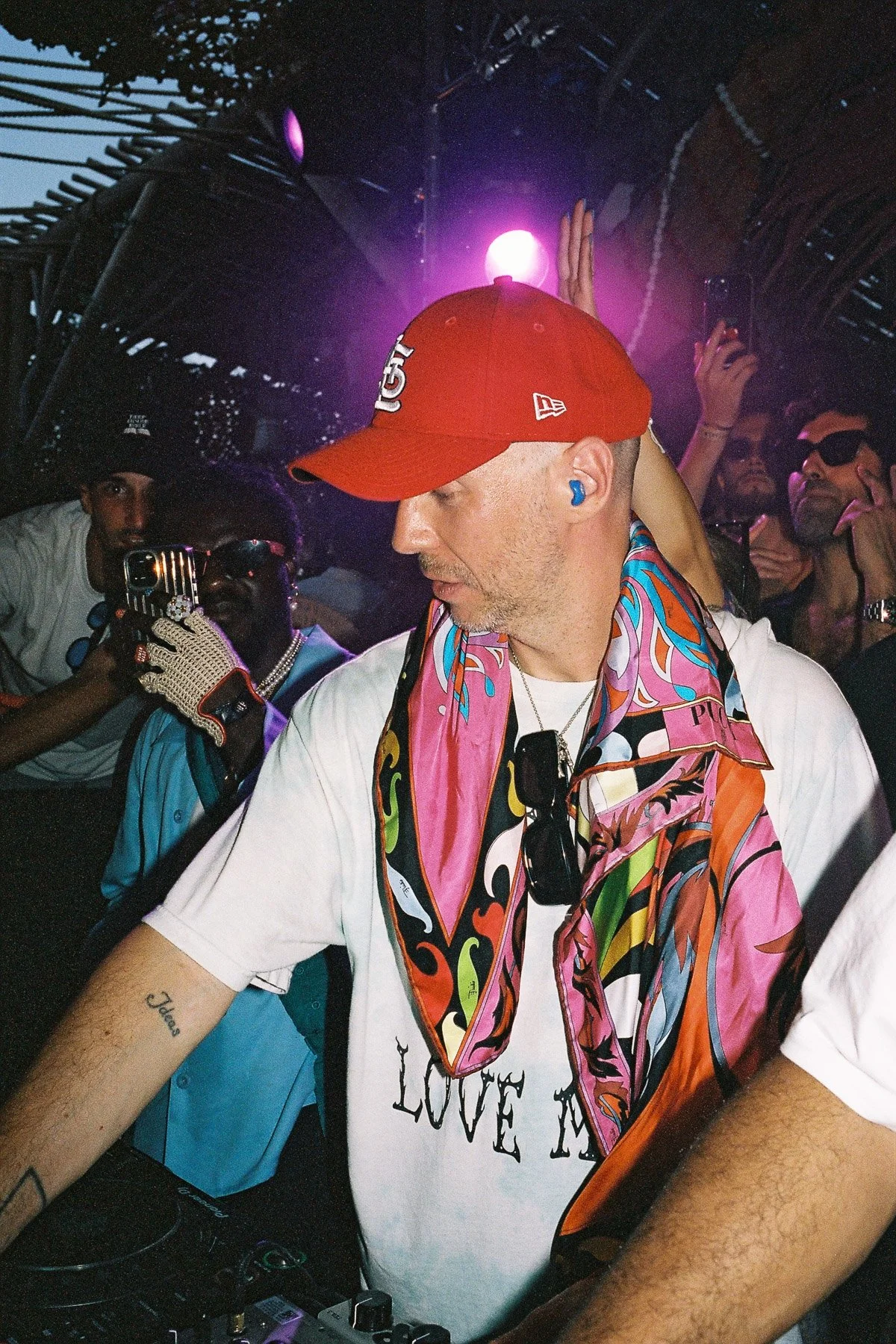 A DJ with a colorful scarf around his neck, a white T-shirt, and a red cap, performing at a crowded party with people around him taking pictures and videos. Purple lights are visible in the background.