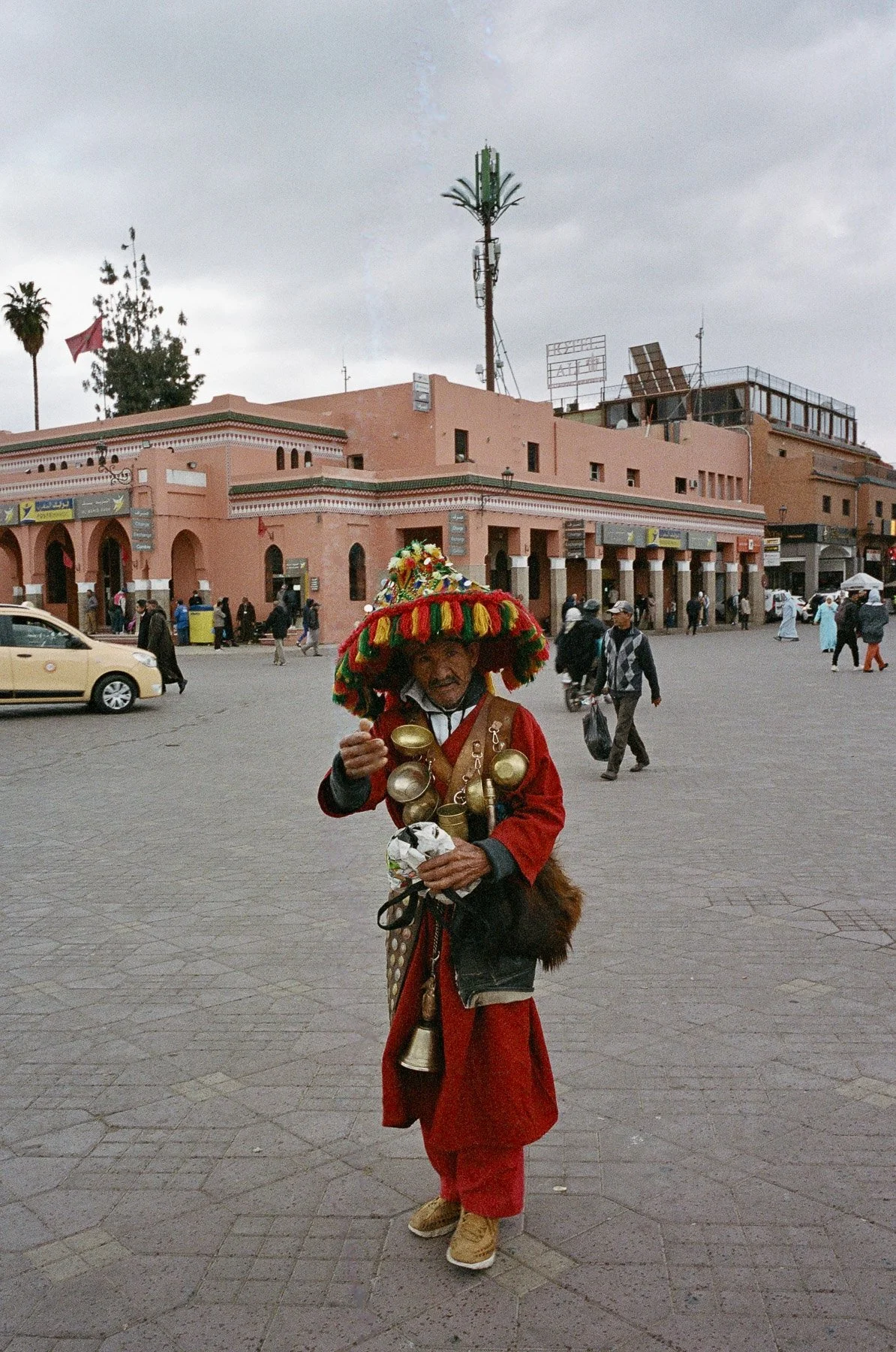 A man dressed in traditional attire stands in a busy plaza, wearing a large colorful hat, with bells and ornaments around his neck, holding a small object in his hand. There are people and a pink building with signs and trees in the background.
