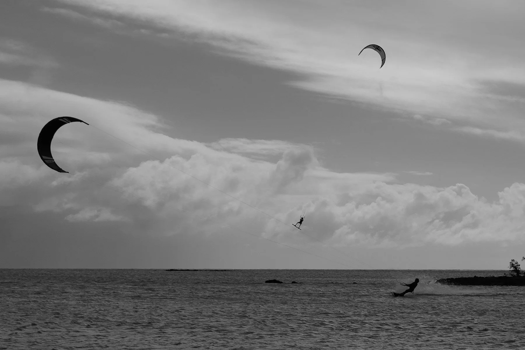 A black and white photograph of a person windsurfing and kite surfing near the shoreline with clouds in the sky.