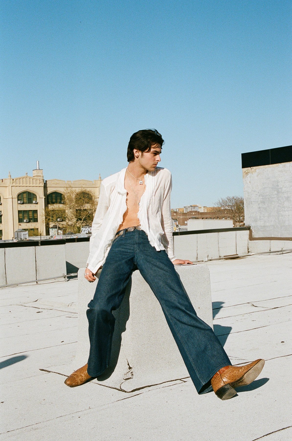 Young man with dark hair, wearing an open white shirt, jeans, and brown shoes, posing on a rooftop during daytime against a clear blue sky and city buildings.