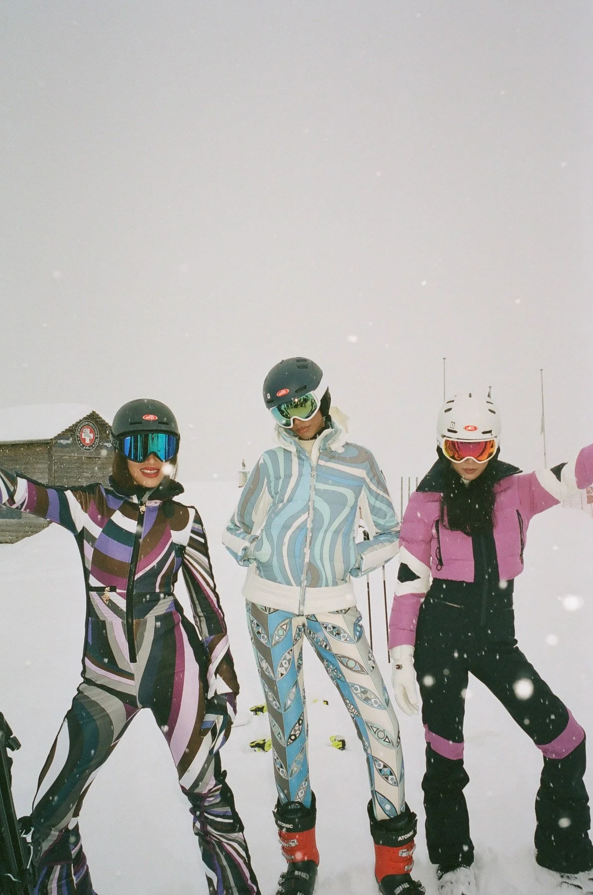Three women dressed in colorful ski outfits wearing helmets and goggles, standing on a snowy ski slope with a ski resort building in the background.