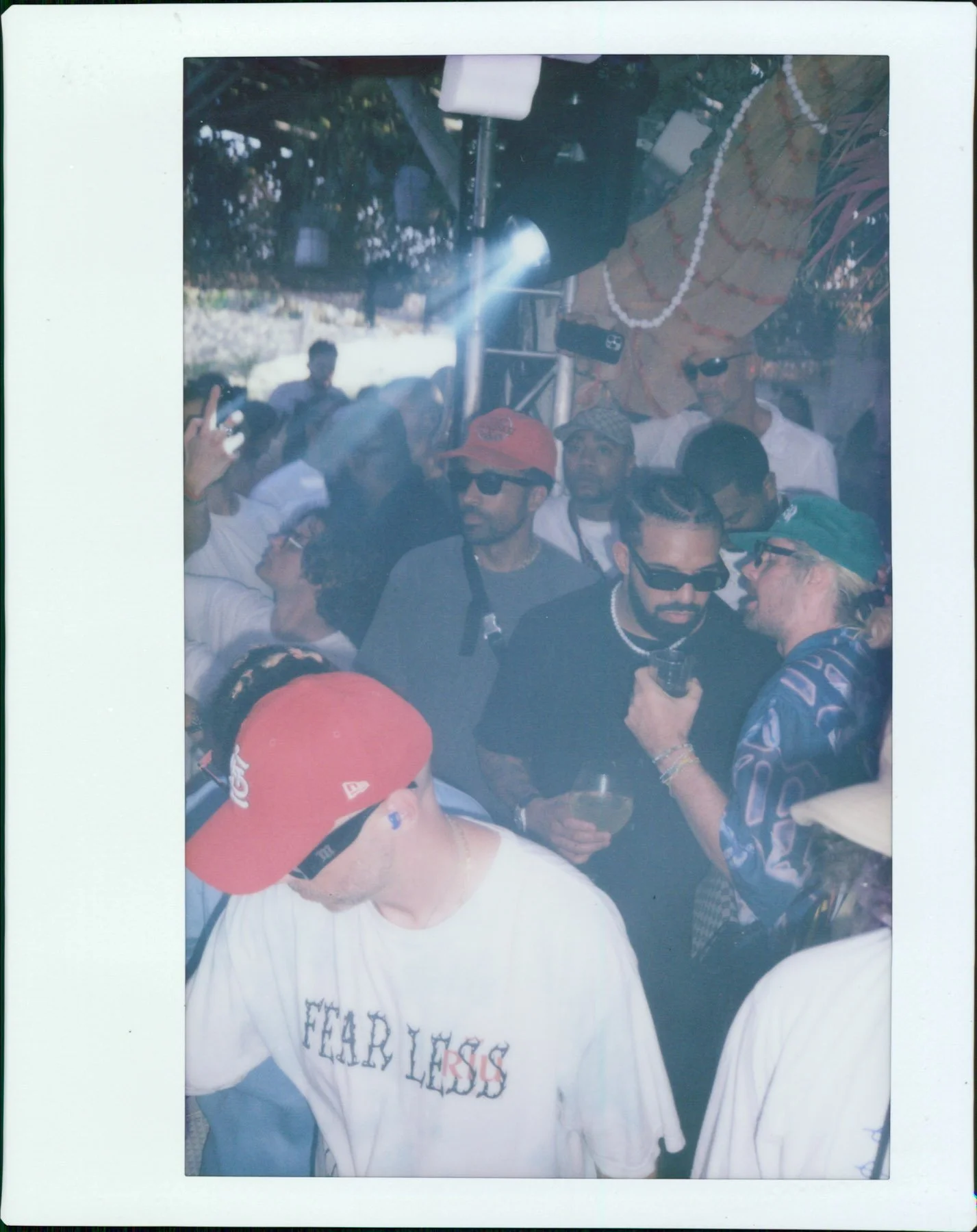 A group of people at an outdoor gathering, some wearing sunglasses and hats, standing under a canopy decorated with flowers and beads. One person is holding a drink, and the scene appears lively with multiple individuals engaged in conversation and e