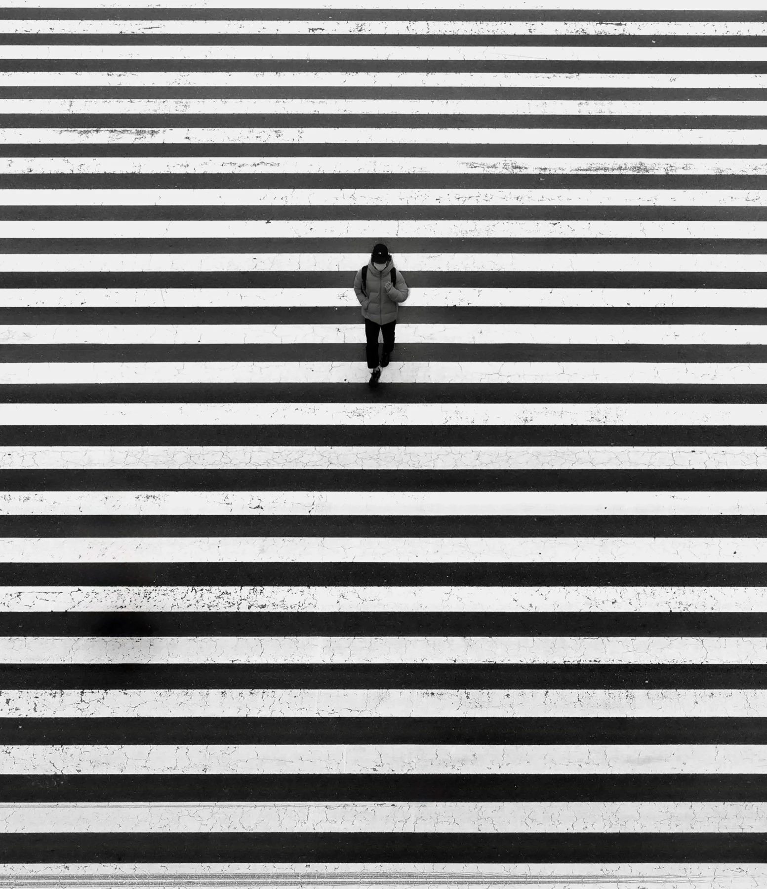 A person walking across a black and white striped crosswalk, wearing a jacket, backpack, and cap, with their head slightly bowed.