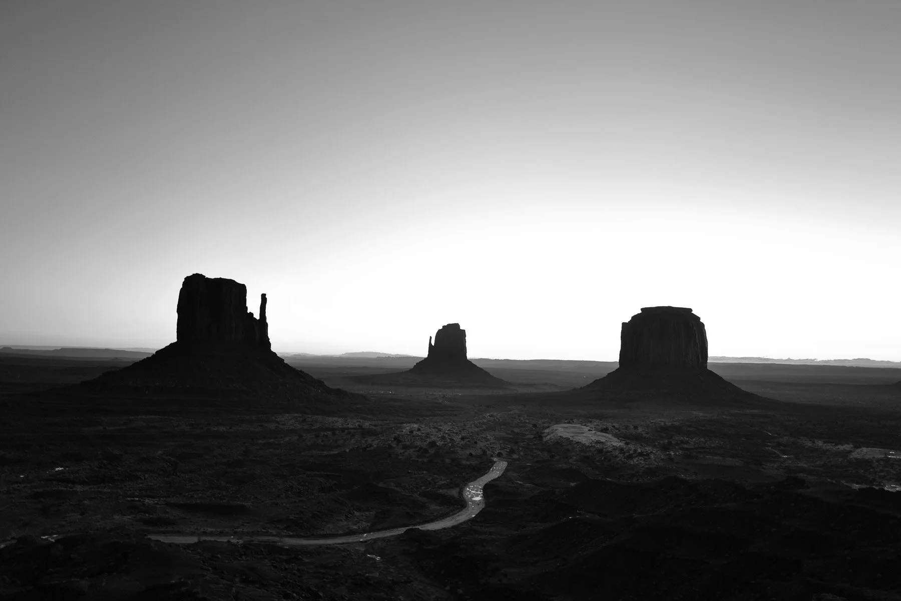 Black and white landscape photo of Monument Valley featuring three large buttes with a winding dirt road in the foreground.