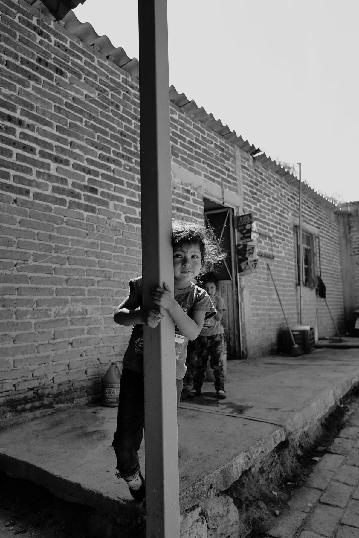 Two children playing outside a brick house, one child peeks around a post, while the other stands with their hands on their hips, in a residential area.