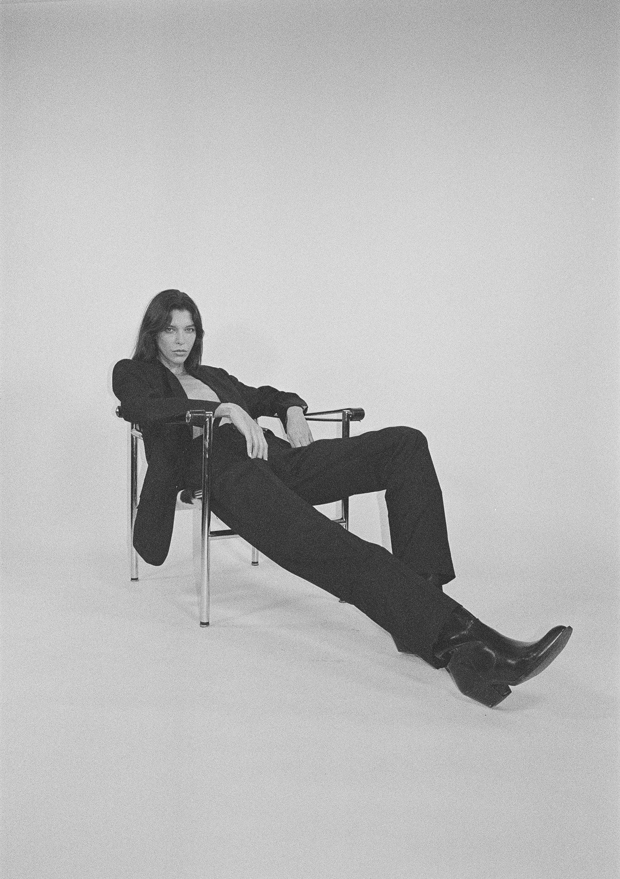 A woman in black clothing and leather boots sitting on a chair with her legs extended forward and looking at the camera, in a minimalistic setting.