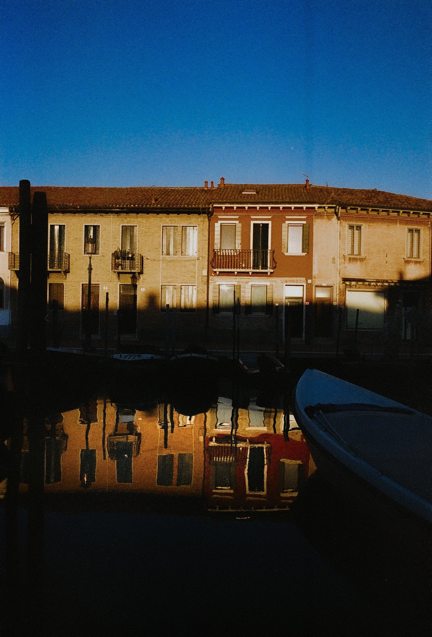 Colorful buildings along a canal with boat reflection, under a clear blue sky at sunset.
