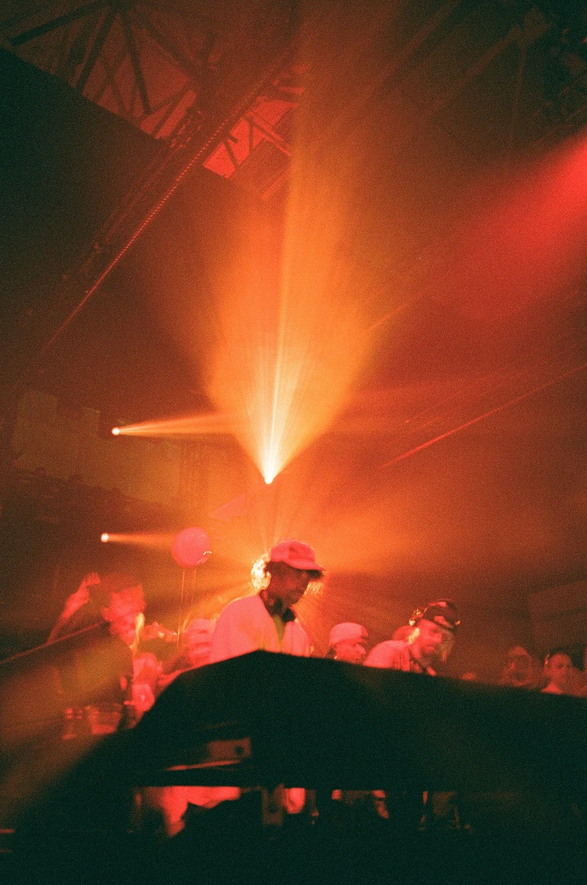 People at a concert or club with vibrant orange and red lighting, some wearing baseball caps, and a visible stage or DJ setup in the foreground.