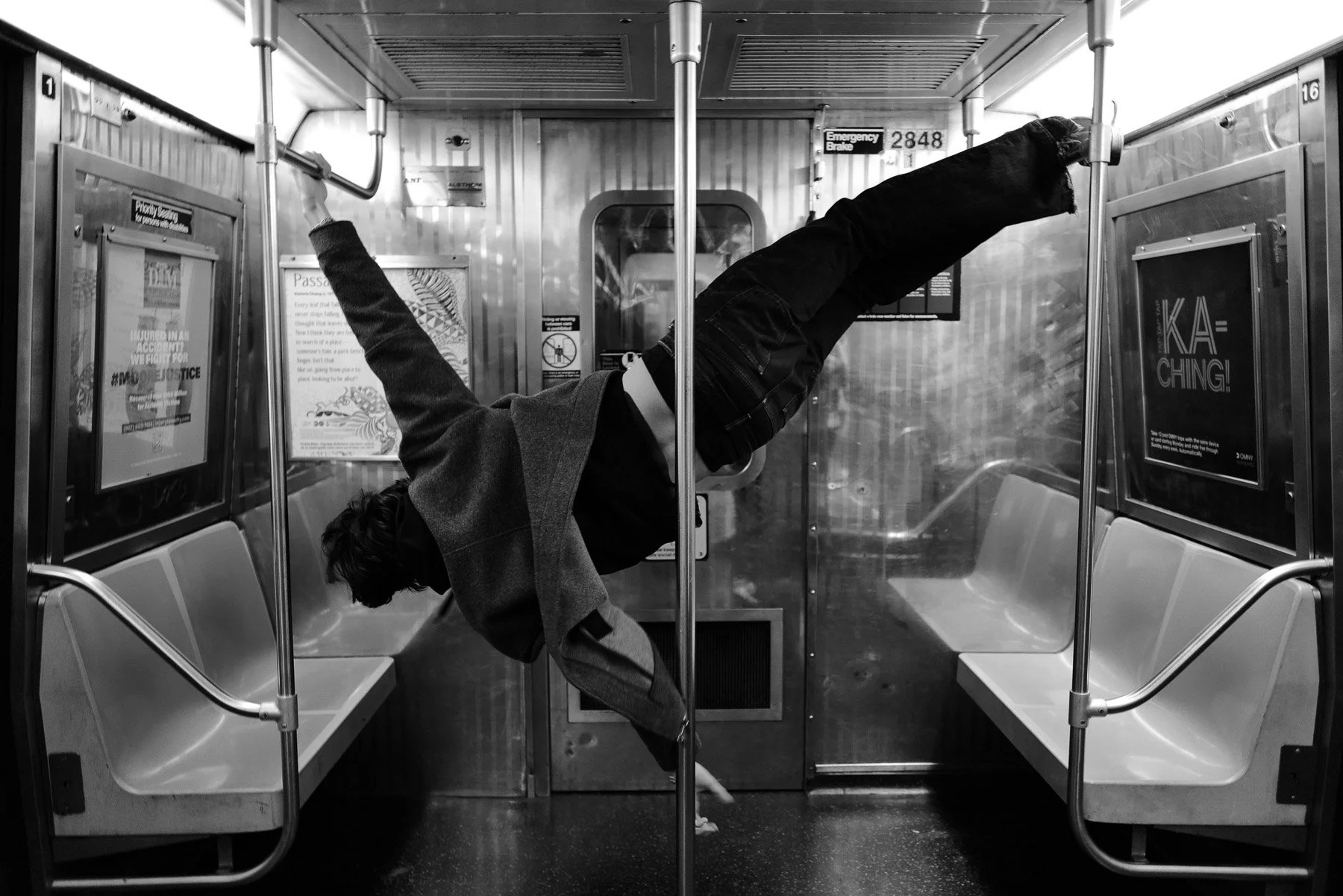 A person dressed in dark clothing is performing a handstand on the aisle of a subway train, with legs extended through the bike of the train. The train is mostly empty, with some empty seats visible and advertisements on the walls.
